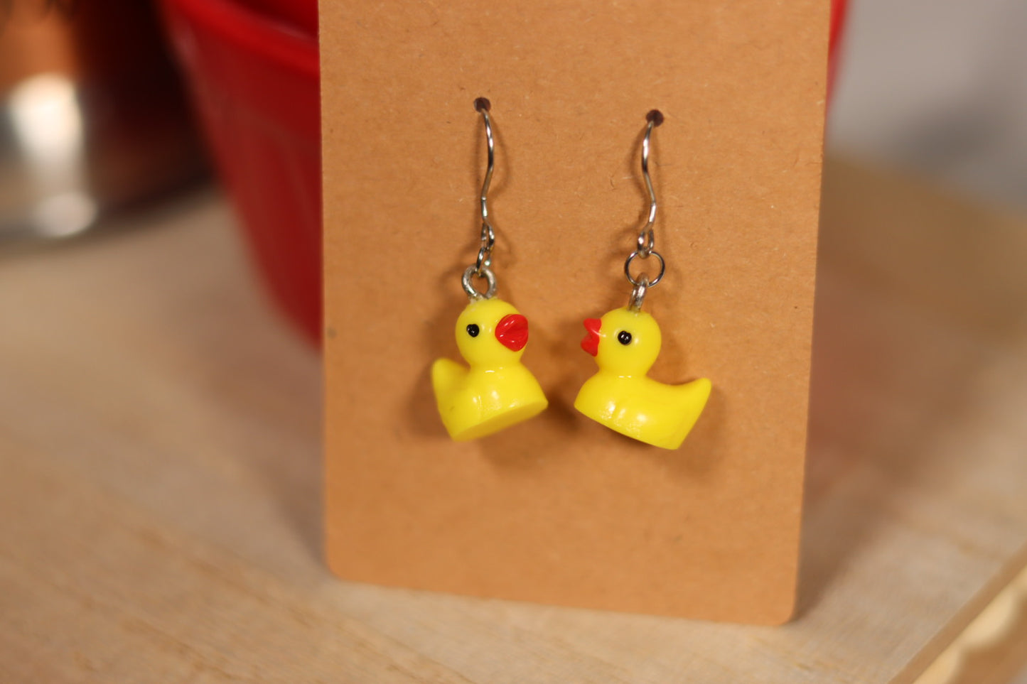 A close-up photograph of a pair of earrings featuring small, yellow rubber duck charms with red beaks and black eyes. The ducks are attached to silver hooks and dangle from a simple brown cardboard backing. The background is slightly blurred, showing a light wooden surface and a red container.