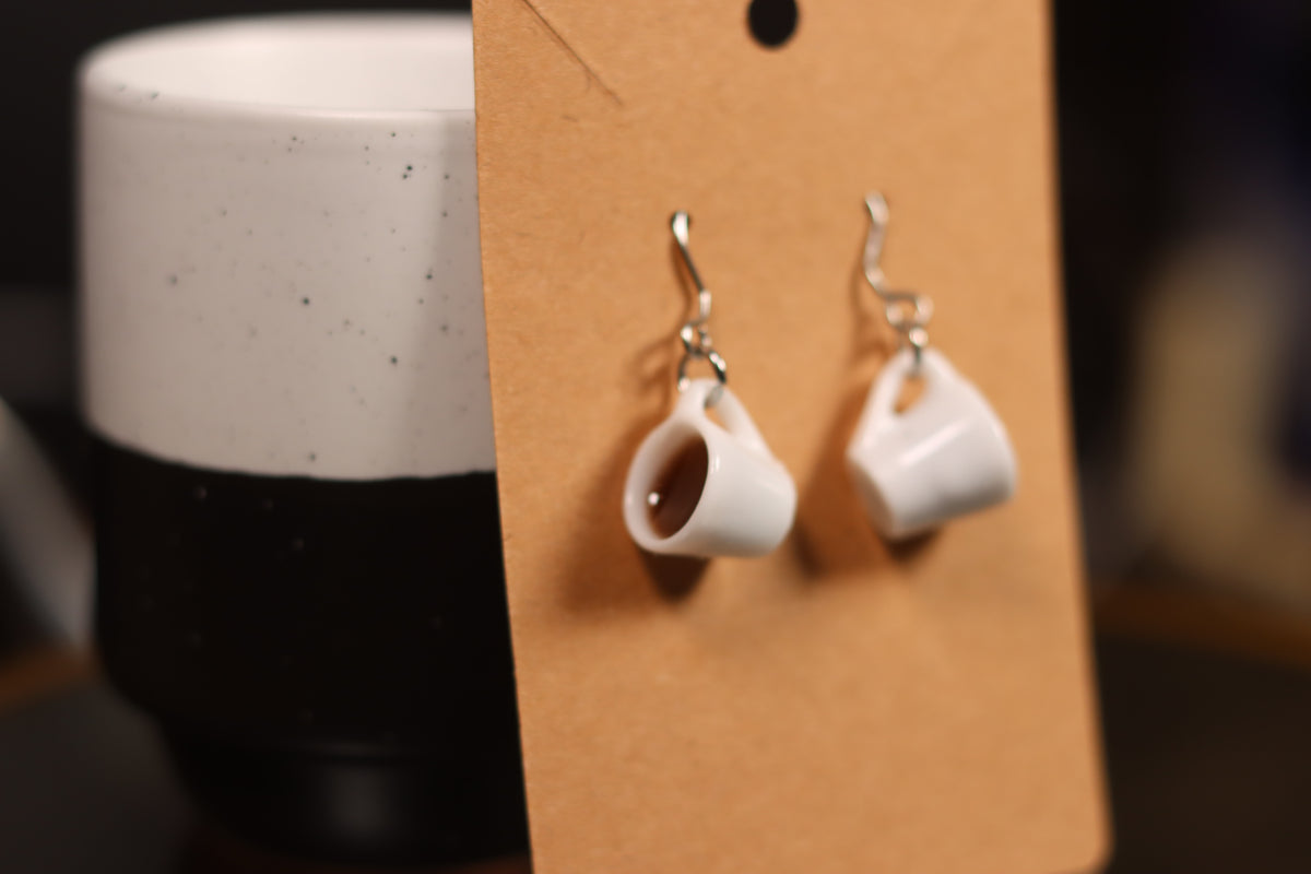 White cup-shaped earrings on a brown card with a blurred coffee cup in the background